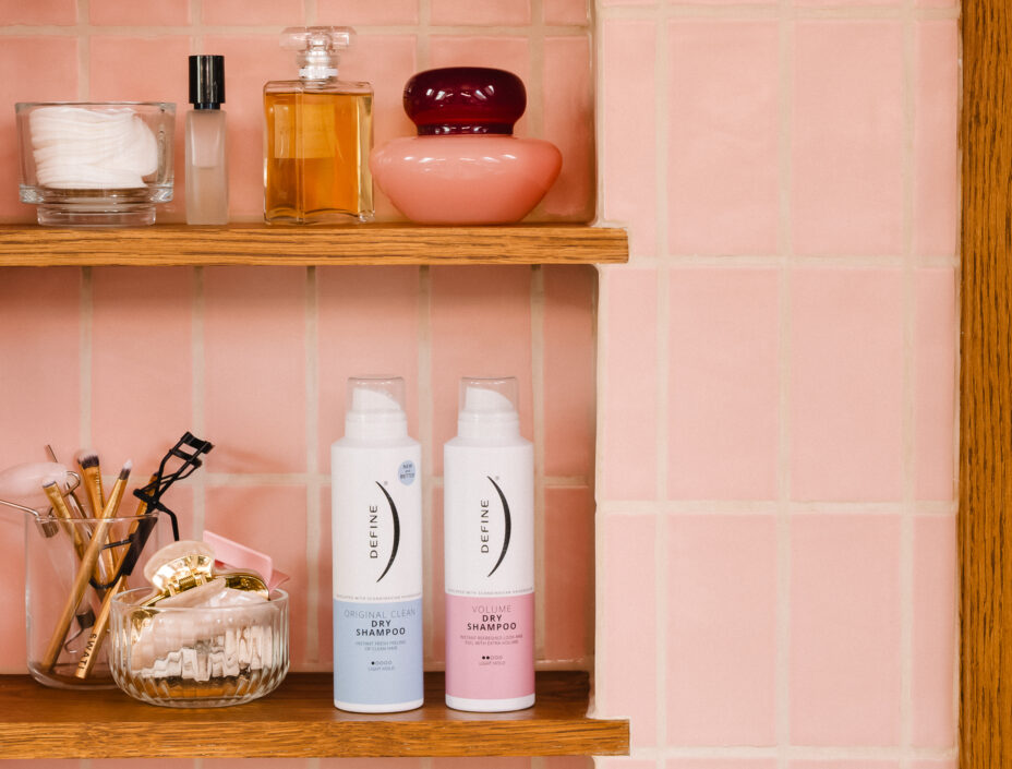 To flasker tørrshampoo står på en baderomshylle bak rosa fliser sammen med parfyme, smykker og baderomspynt