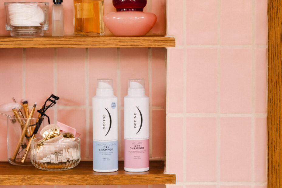 To flasker tørrshampoo står på en baderomshylle bak rosa fliser sammen med parfyme, smykker og baderomspynt