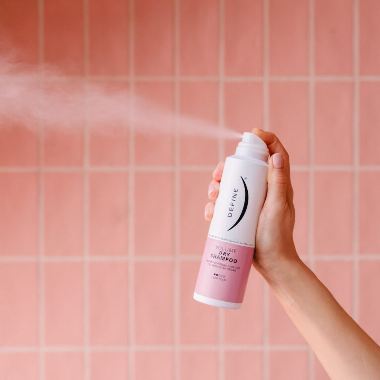 Tørrshampoo flaske bak rosa fliser som sprayer produkt ut i en hvit sky i lufta